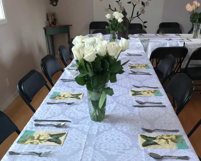 A long table is set with white tablecloths, black chairs, and a vase of white roses at the center, surrounded by plates, utensils, and napkins.