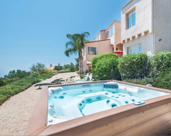A hot tub sits in a landscaped yard with a view of a house and surrounding greenery.