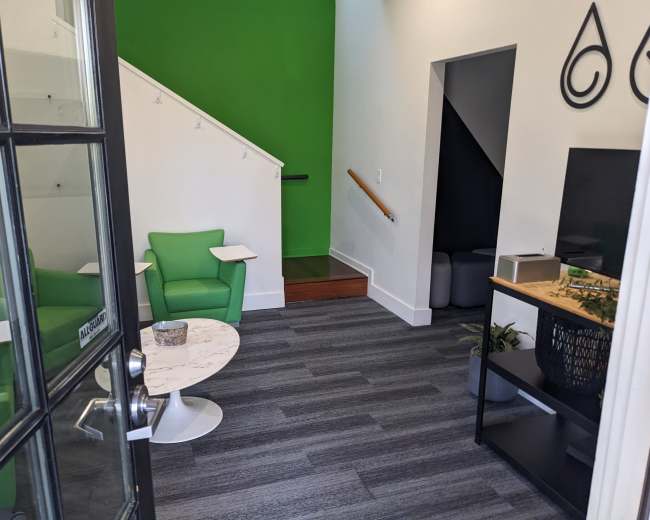 The image shows a modern interior space with green walls, a small seating area featuring green chairs, a marble-top table, and a staircase leading to an upper level.