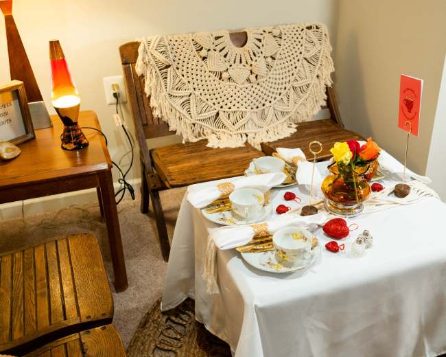 A small table is set for tea with fine china, a vase of roses, and decorative items, surrounded by a crocheted shawl and wooden furniture.