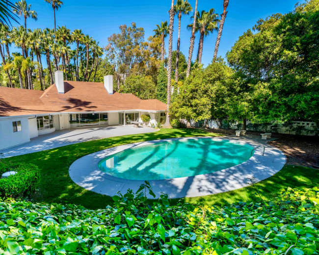 The image shows a backyard with a swimming pool surrounded by lush greenery and palm trees, adjacent to a single-story house.