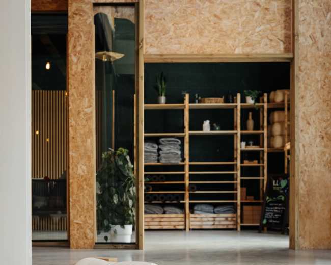 The image shows a yoga studio with mats laid out on a smooth floor, featuring props like bolsters and blocks, and shelving in the background displaying stacked towels.