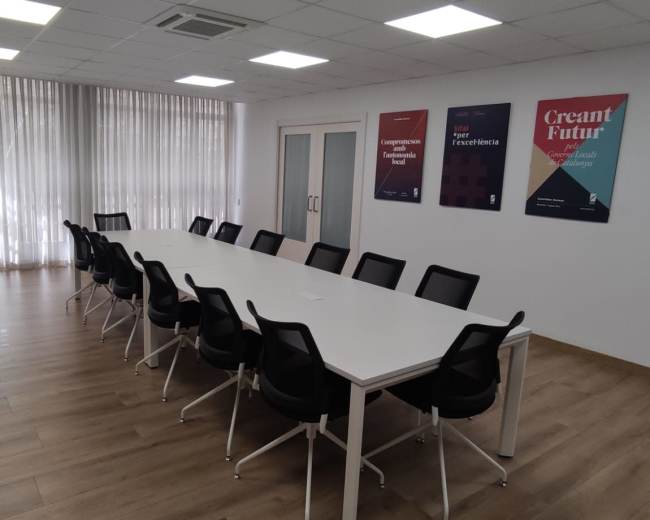 A well-lit conference room features a long white table surrounded by black chairs, with shelves and posters on the walls.