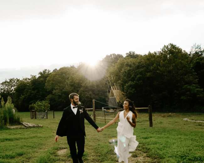 A couple in formal attire walks hand in hand down a dirt path in a rural setting with trees in the background and sunlight peeking through the clouds.