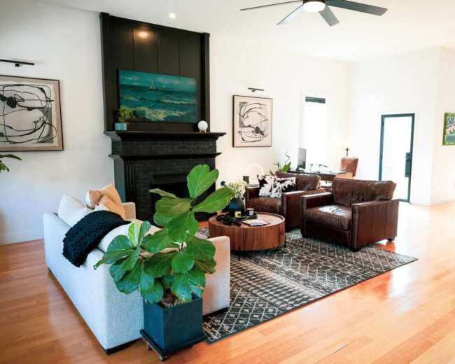 The image shows a modern living room with a white sofa, brown leather armchairs, a round coffee table, and a green plant, all arranged around a patterned rug and a black fireplace.