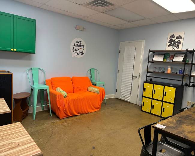 The image shows a small, brightly colored room featuring an orange couch, two teal chairs, a black shelf with yellow lockers, and a wall with a decorative sign.