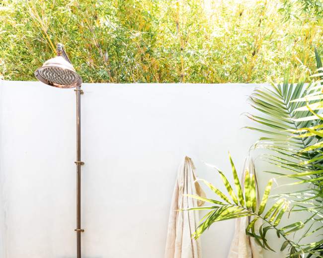 The image features an outdoor shower made of copper piping, with two light-colored towels hanging nearby and a wooden stool on a wooden deck.