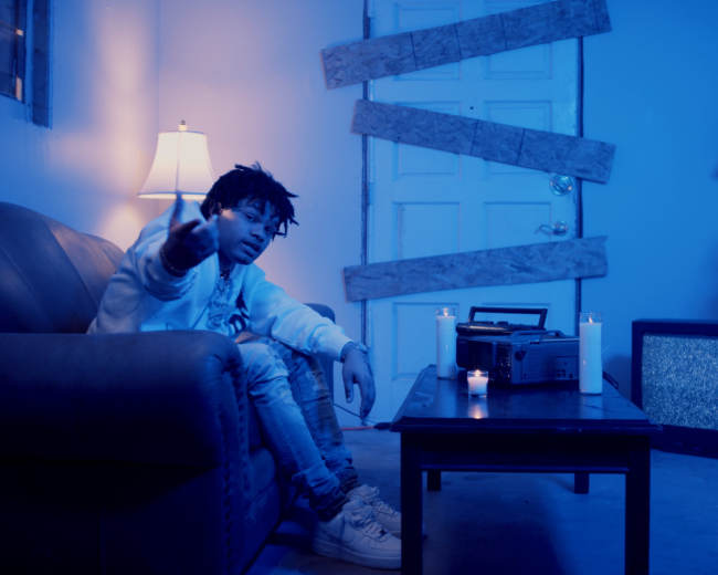 A person sits on a couch in a dimly lit room with a lamp, candles, and a recording device on a table, while wooden planks cover the door.