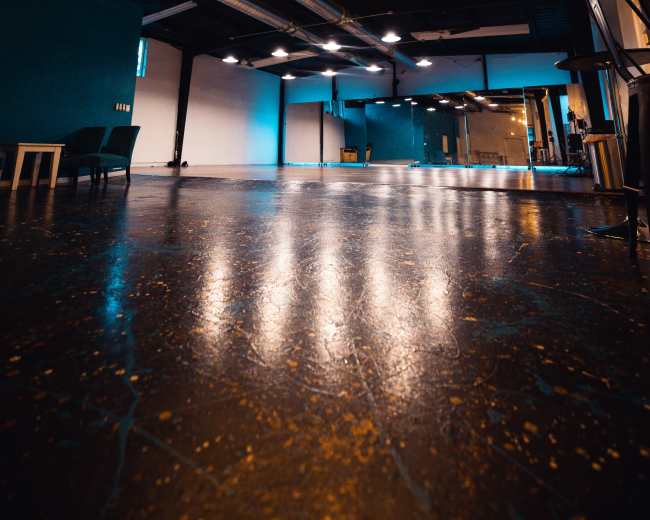 The image shows a spacious, empty room with a polished floor, industrial lighting, and a large chandelier hanging from the ceiling.