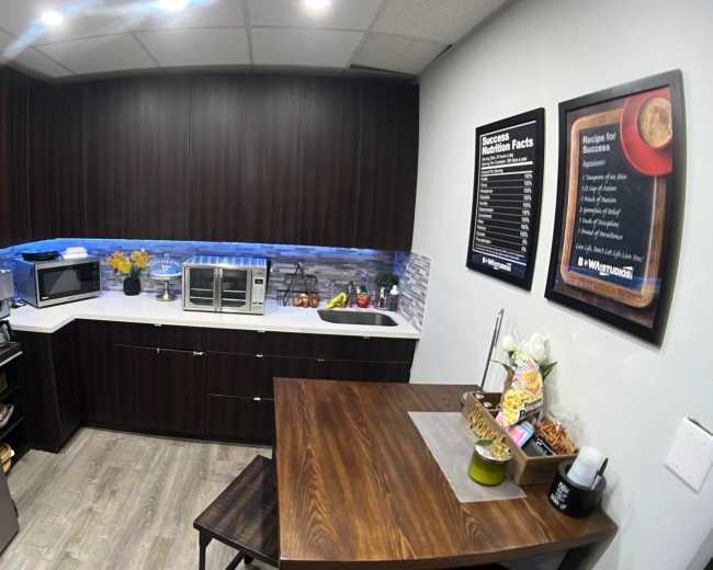 The image shows a modern kitchen with dark cabinetry, a stainless steel refrigerator, a microwave, a toaster oven, and a small dining table.