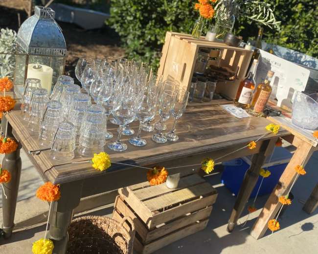 A wooden table is set up with an arrangement of clear glasses, bottles of whiskey, and decorative marigold flowers.