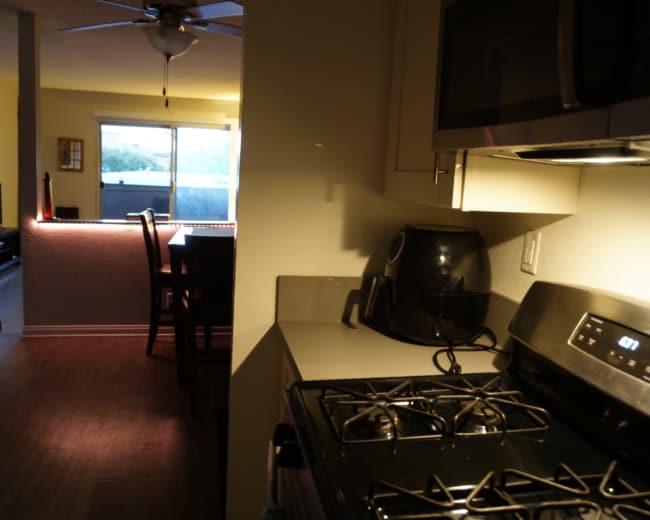 The image shows a kitchen with a gas stove and microwave, adjacent to a dining area illuminated by artificial light.