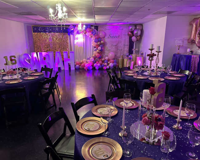 A decorated event space for a birthday celebration, featuring tables set with plates and glasses, a large "16" display, and colorful balloon arrangements.