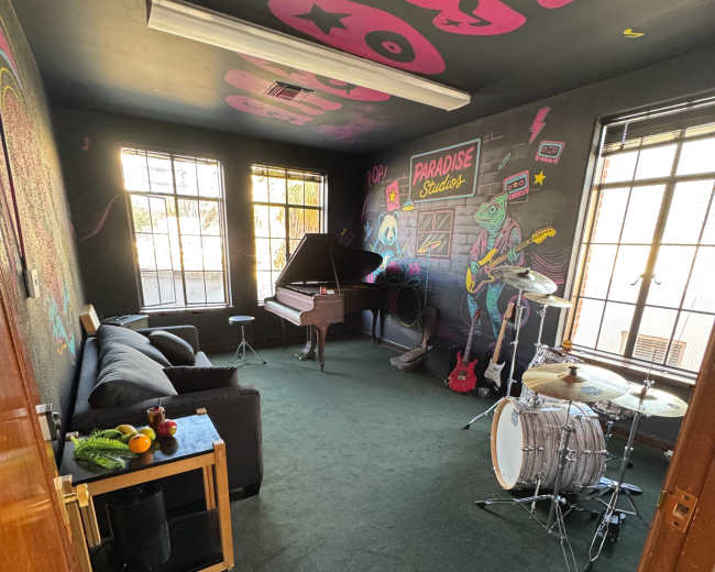 The image shows a music studio with a grand piano, a drum set, a guitar, a couch, and colorful wall murals.
