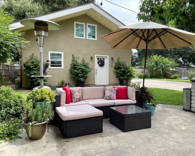 The image shows an outdoor seating area with a sectional sofa adorned with pink pillows, a patio umbrella, a gas heater, and a variety of potted plants, all set in front of a small house with a landscaped yard.
