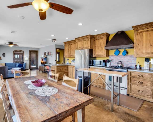 The image shows a modern kitchen with wooden cabinets and a large dining table, adjacent to a living area with a blue couch visible in the background.