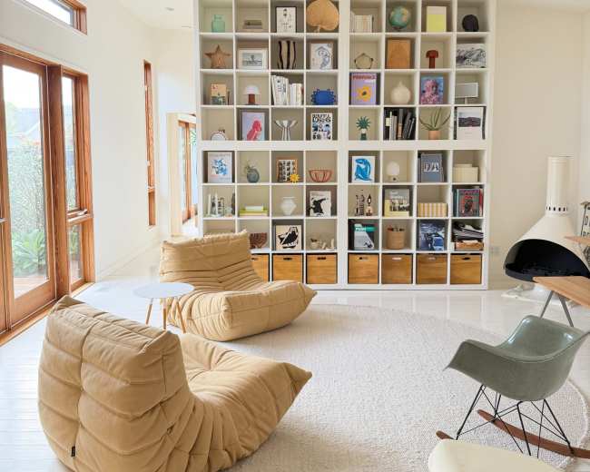 The image shows a modern living room featuring a large bookshelf filled with various decorative items and books, two padded chairs, a round rug, and large windows.