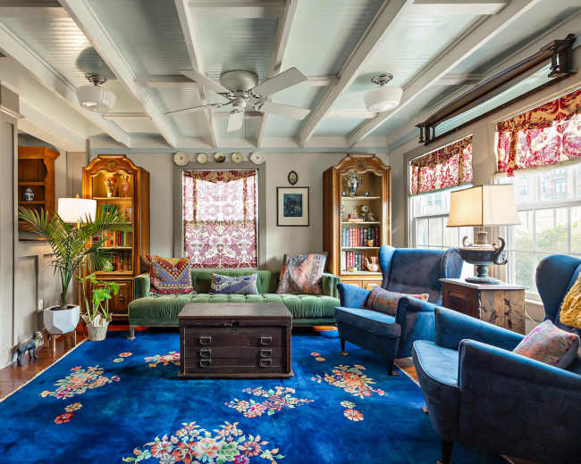 The image shows a cozy living room with a green sofa, two blue chairs, a dark wooden coffee table, and bookshelves filled with books and decorative items.
