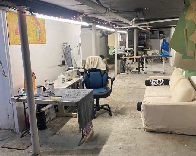 The image shows a spacious basement area with a sewing station, a blue office chair, a beige couch, and various art supplies scattered around.