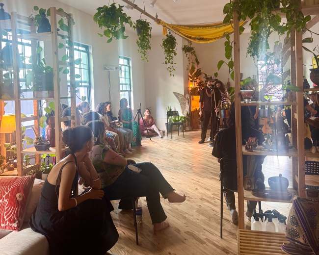 A spacious indoor venue is filled with people seated among plants and decorative items, while a performer stands at the front.