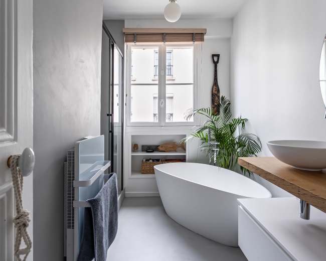 A modern bathroom features a freestanding tub, a wooden counter with a bowl sink, and large windows allowing natural light, complemented by indoor plants.