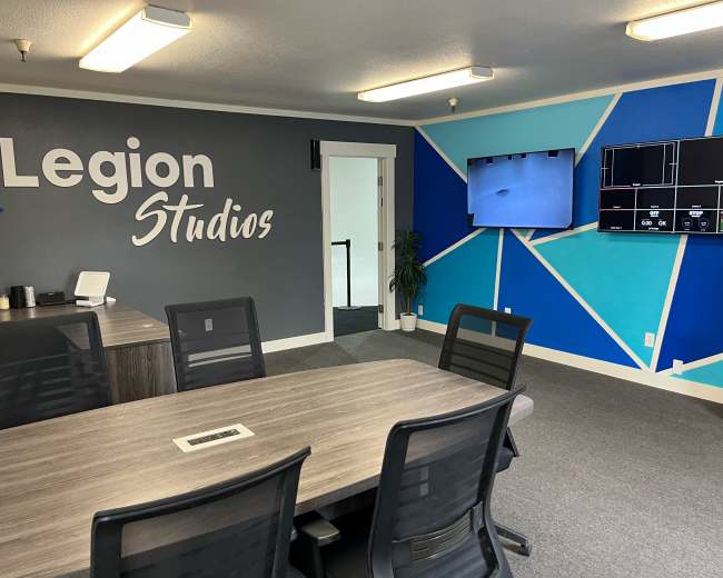 The image shows a modern office space featuring a large conference table surrounded by black chairs, a wall with the text "Legion Studios," and a screen displaying digital content.