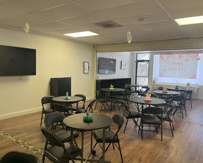 The image shows a decorated room with black circular tables and chairs arranged for a birthday celebration, featuring a "Happy Birthday" banner in the window.