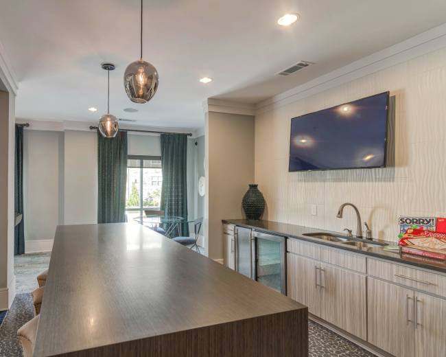 The image shows a modern kitchen space with a long countertop, pendant lights, a flat-screen TV mounted on the wall, and a small bar area containing snacks.