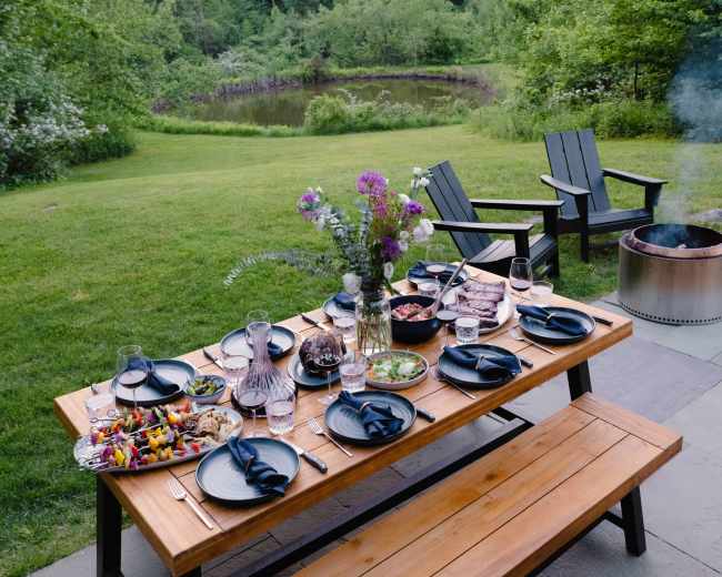A long wooden table is set with plates and glasses, surrounded by a lush green landscape and a tranquil pond, with two Adirondack chairs nearby.