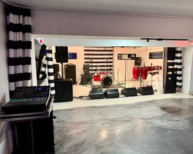 The image shows a music performance space featuring a drum set, congas, amplifiers, and sound equipment set against a backdrop of black and white striped decor.