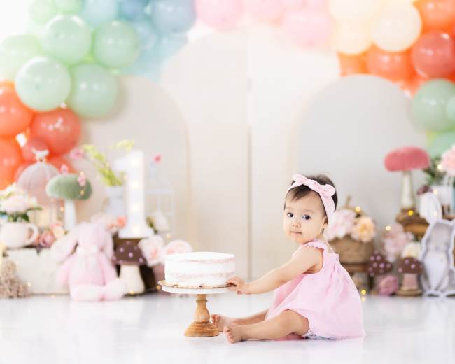 A baby in a pink dress sits on the floor while holding a small cake, surrounded by colorful balloon decorations and stuffed animals.