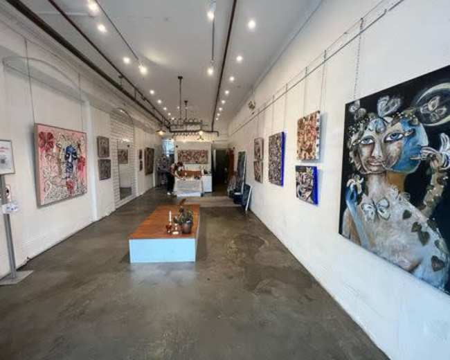 The image shows a modern art gallery with paintings displayed on the white walls and a wooden table in the center of the space.