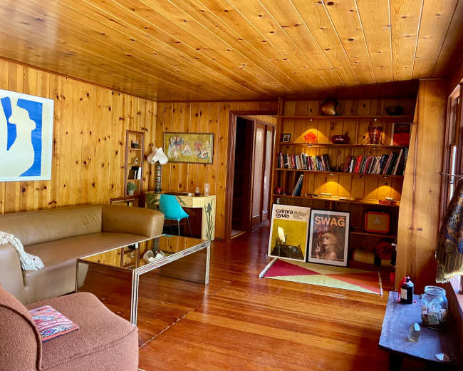 The image shows a wooden-paneled living room with a sofa, a glass coffee table, a bookshelf filled with books, and artwork on the walls.