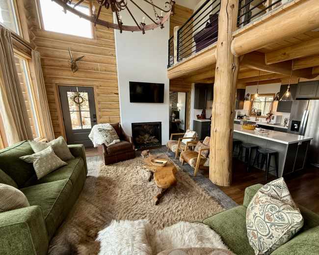 The image shows a cozy living room with a large fireplace, green sofas, and a view of a kitchen area featuring modern appliances and wooden accents.
