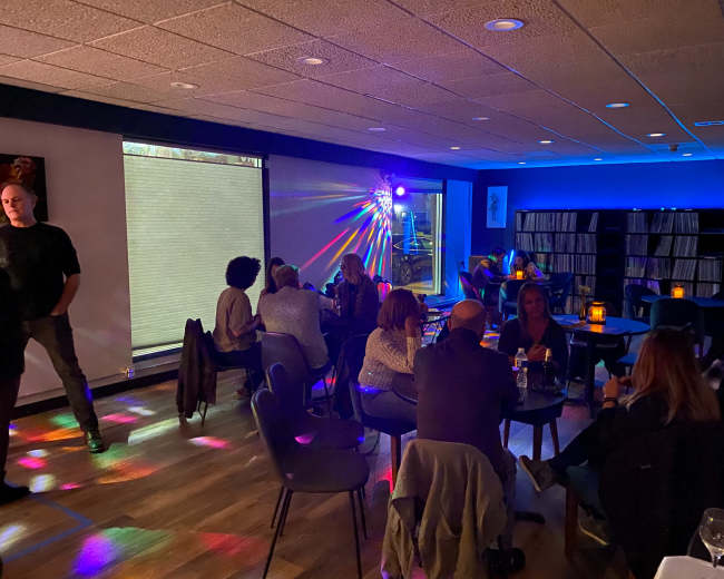 A dimly lit lounge features patrons seated at tables while colorful lights create patterns on the floor and walls.