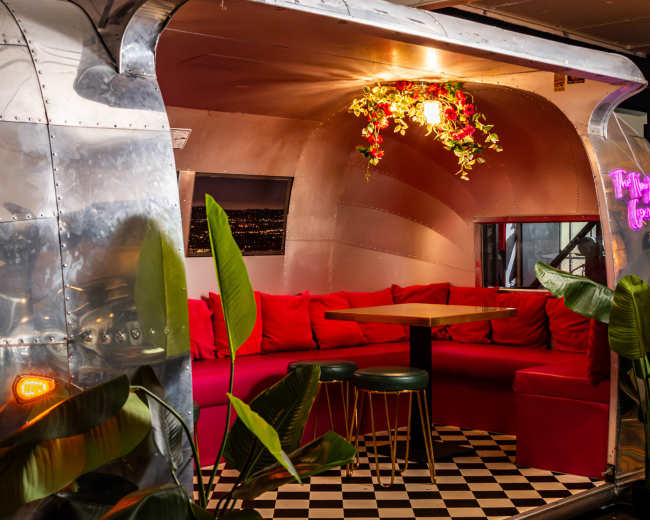 The interior features a rounded, metallic booth adorned with red seating and a small table, surrounded by green plants and a checkered floor.