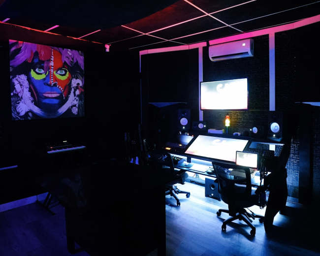 A dark music studio features colorful lighting, a large monitor, and professional audio equipment.