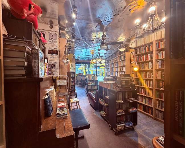 The image shows the interior of a cozy bookshop with shelves filled with books, wooden furniture, and warm lighting.