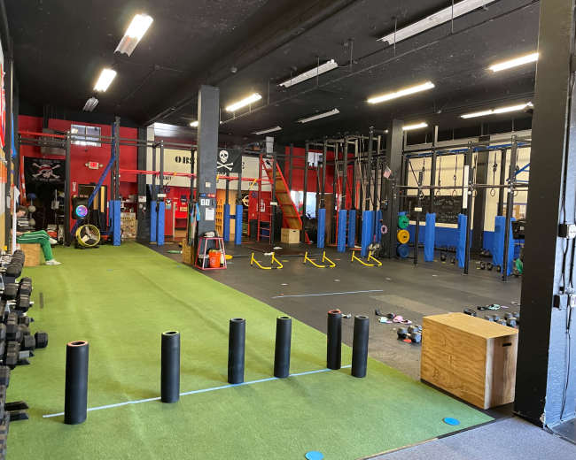 The image shows a well-equipped gym with various fitness stations, including weights, pull-up bars, and exercise mats, set up on a green turf floor.