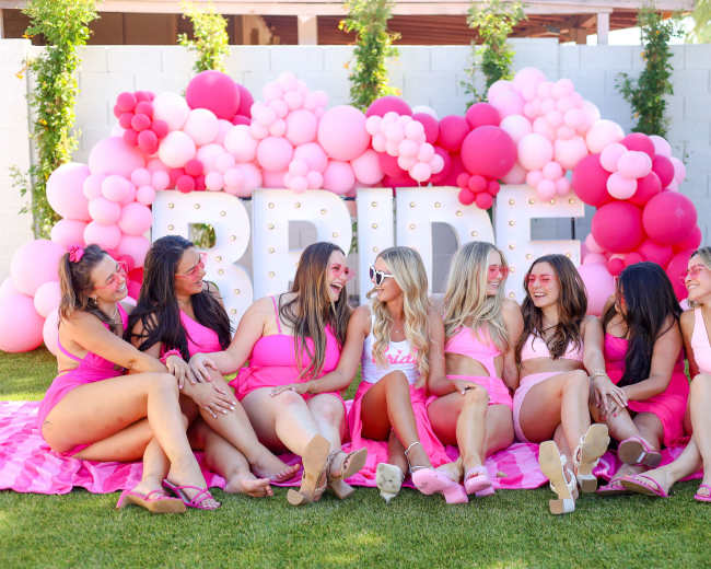 A group of women in pink swimsuits sit together on a blanket in front of a balloon arch that spells "BRIDE."
