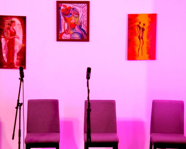 A stage setup with three empty chairs and two microphones, framed by colorful artwork on a pink-lit wall.