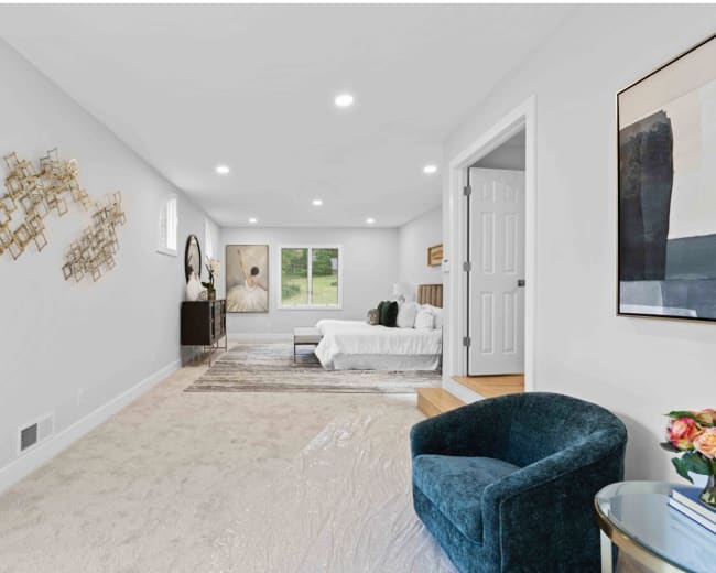The image shows a spacious, light-colored bedroom with a bed in the back, a decorative chair in the foreground, and large windows providing natural light.