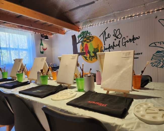 The image shows a painting setup with empty canvases, paintbrushes, and palettes arranged on a table in a colorful, artistically decorated room.
