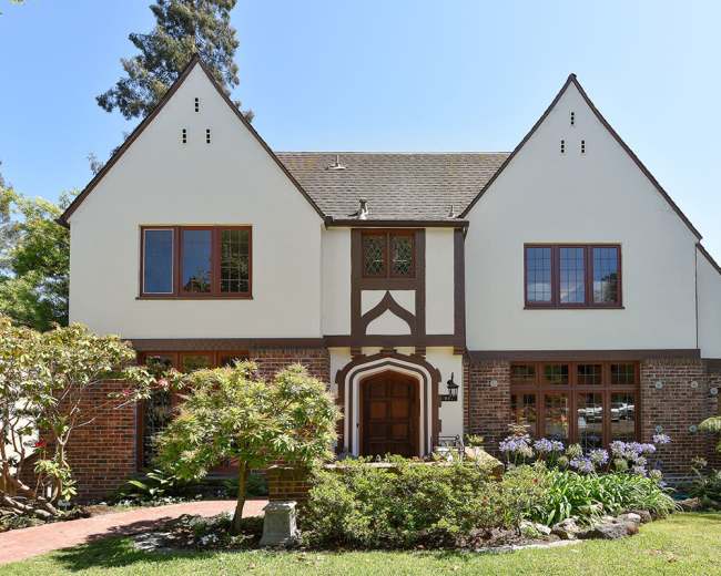 The image shows a two-story Tudor-style house with a brick base, white stucco upper walls, and a landscaped front yard featuring various plants and flowers.