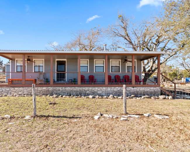 The image shows a single-story house with a covered porch, featuring several red chairs and surrounded by a grassy area and sparse trees.