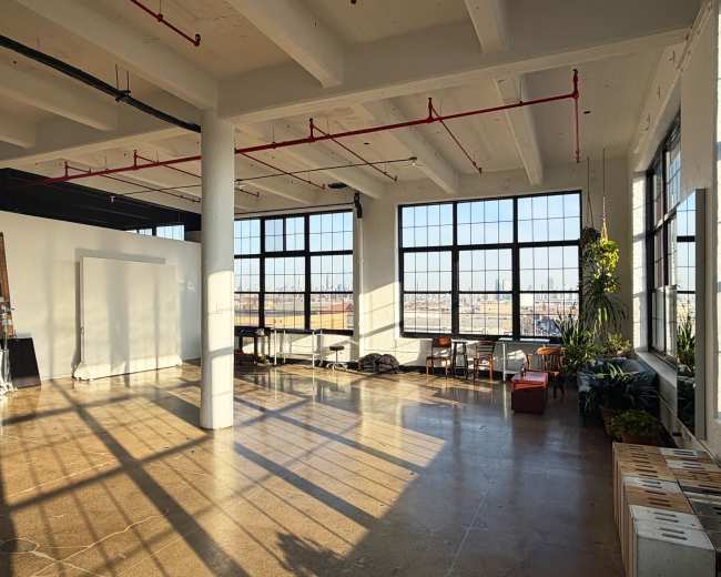 The interior of a spacious, sunlit room features large windows, polished concrete floors, and a few scattered pieces of furniture.