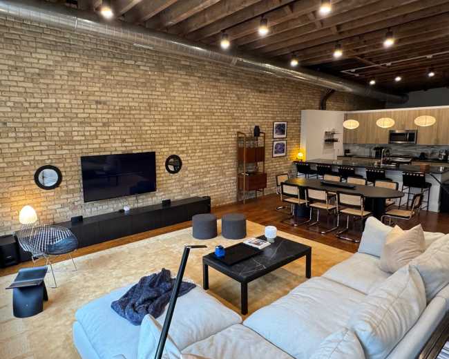 The image shows a modern living room with exposed brick walls, a large sectional sofa, a coffee table, and an open kitchen area with dining seating in the background.