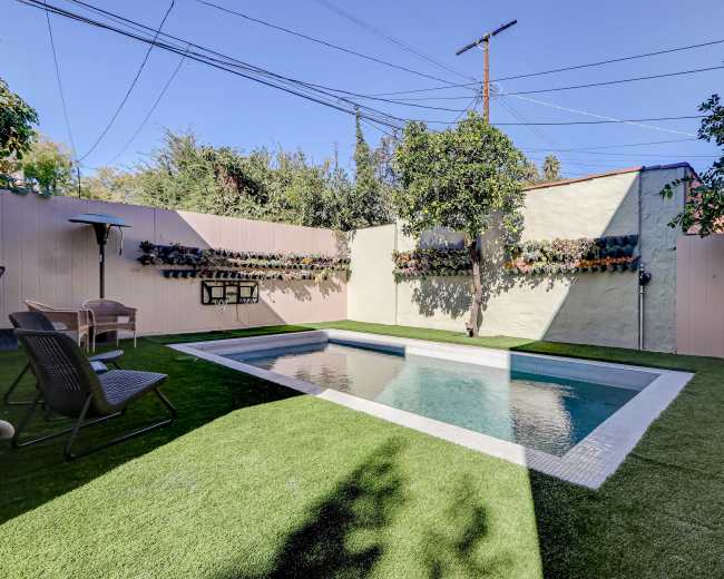 The image shows a small, rectangular swimming pool surrounded by artificial turf and bordered by walls with greenery and outdoor seating.