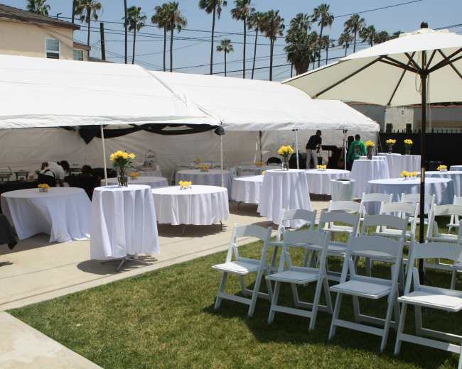 A tented outdoor area is set up for an event, featuring round tables with white tablecloths, chairs, and decorations, surrounded by palm trees.