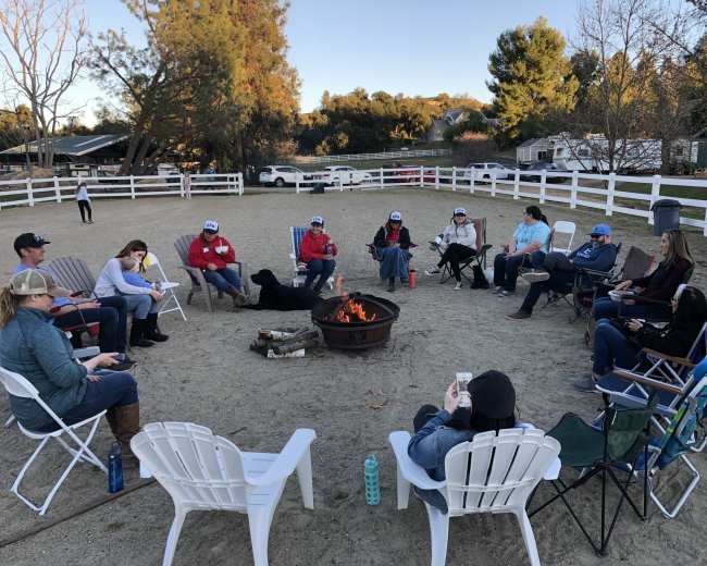 A group of people sits in chairs around a fire pit in a sandy area, with trees and a white fence in the background.